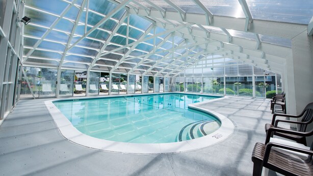 Une piscine intérieure dotée d’un toit en verre, avec une eau cristalline et quelques chaises disposées le long des bords.