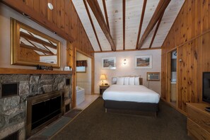 A bedroom with a stone fireplace, a bed with white linens, wooden walls, and a ceiling with exposed beams. - Adirondack Retreat Inn & Suites (Lake George)