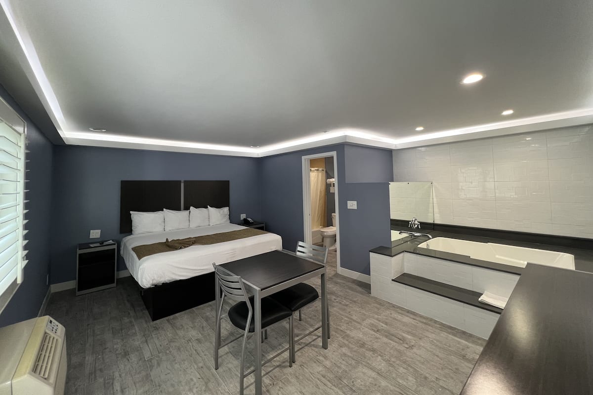 A modern hotel room with a bed, bedside table, chair, and a bathroom with a bathtub and sink.