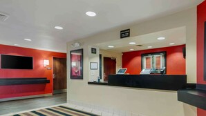 Un espace d’accueil d’hôtel aux couleurs rouge et beige, avec un comptoir d’accueil, un écran numérique et un panneau indiquant les heures d’ouverture.