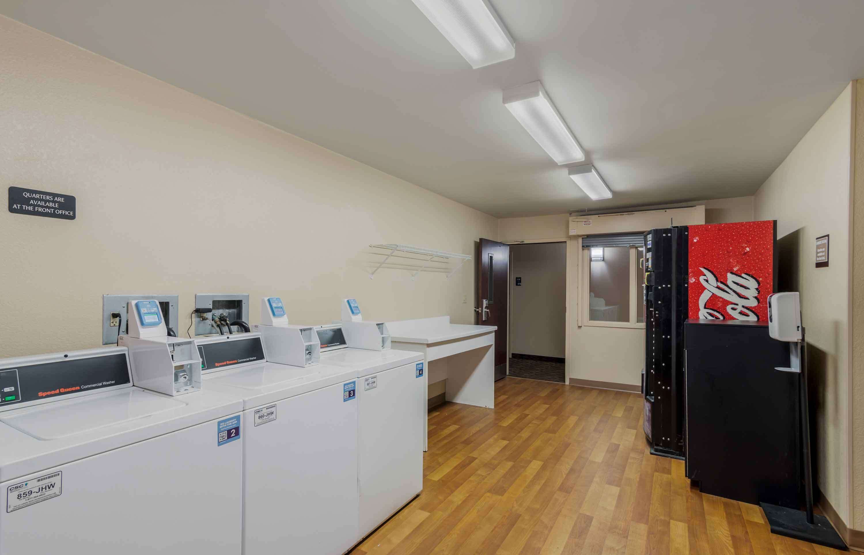 A laundry area with commercial washers and dryers, a Coca-Cola refrigerator, and a sign indicating quarters are available at the front office.