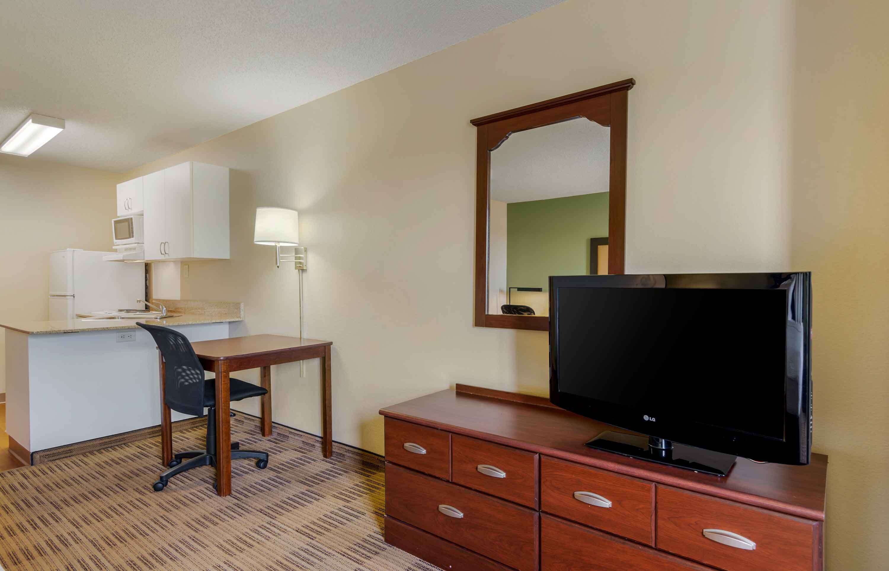 A hotel room with a kitchenette, a desk with a chair, a television on a wooden dresser, and a mirror on the wall.