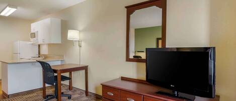 A hotel room with a kitchenette, a desk with a chair, a television on a wooden dresser, and a mirror on the wall.