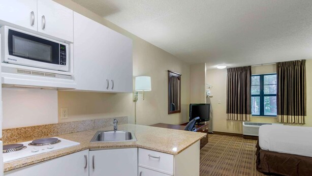 Une chambre d’hôtel compacte dotée d’une kitchenette comprenant un four à micro-ondes, une cuisinière et un évier. La chambre comprend un lit, un téléviseur et une fenêtre avec des rideaux.