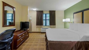 Une chambre d’hôtel comprenant un lit, un bureau avec une télévision, une chaise, une commode, une fenêtre avec des rideaux et une lampe fixée au mur.