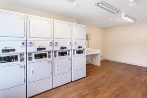 A laundry room with multiple white washing machines, a white countertop, and a wooden floor. - HomeTowne Studios By Red Roof Colorado Springs - Airport (Colorado Springs)