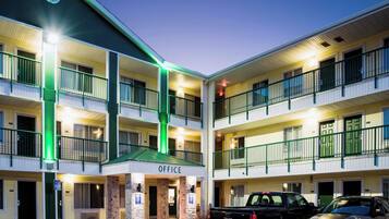 Un hotel de varios pisos con una entrada principal para oficinas, varios balcones y un estacionamiento con dos vehículos.
