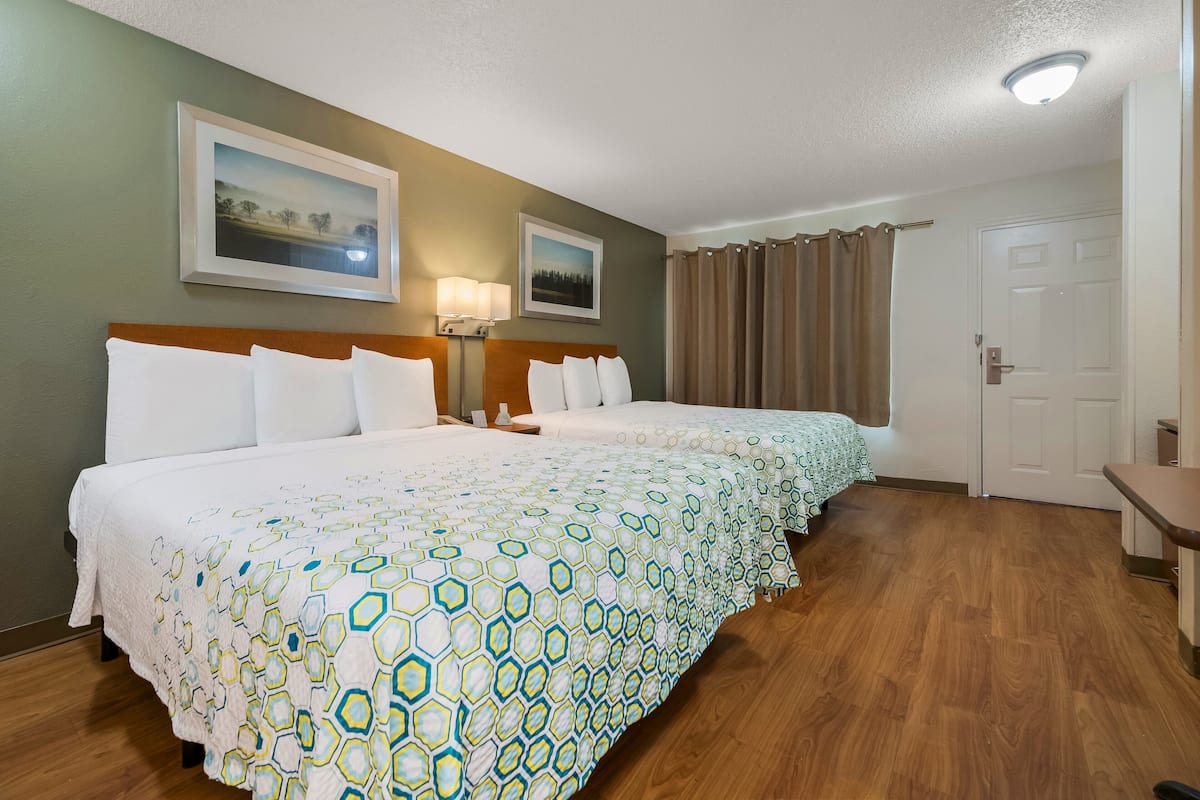 A hotel room with two beds, a wooden floor, a white door, and two framed pictures on the wall.