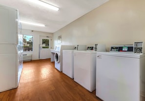 A laundry room with multiple washing machines and dryers, wooden flooring, and a view through a door. - HomeTowne Studios by Red Roof Orlando - UCF Area (Orlando)