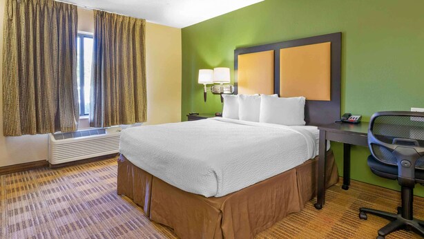 A hotel room with a large bed, a desk with a chair, a window with curtains, and a wall-mounted lamp. - Extended Stay America Suites Jacksonville Camp Lejeune (Jacksonville)