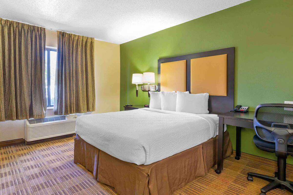 A hotel room with a large bed, a desk with a chair, a window with curtains, and a wall-mounted lamp.