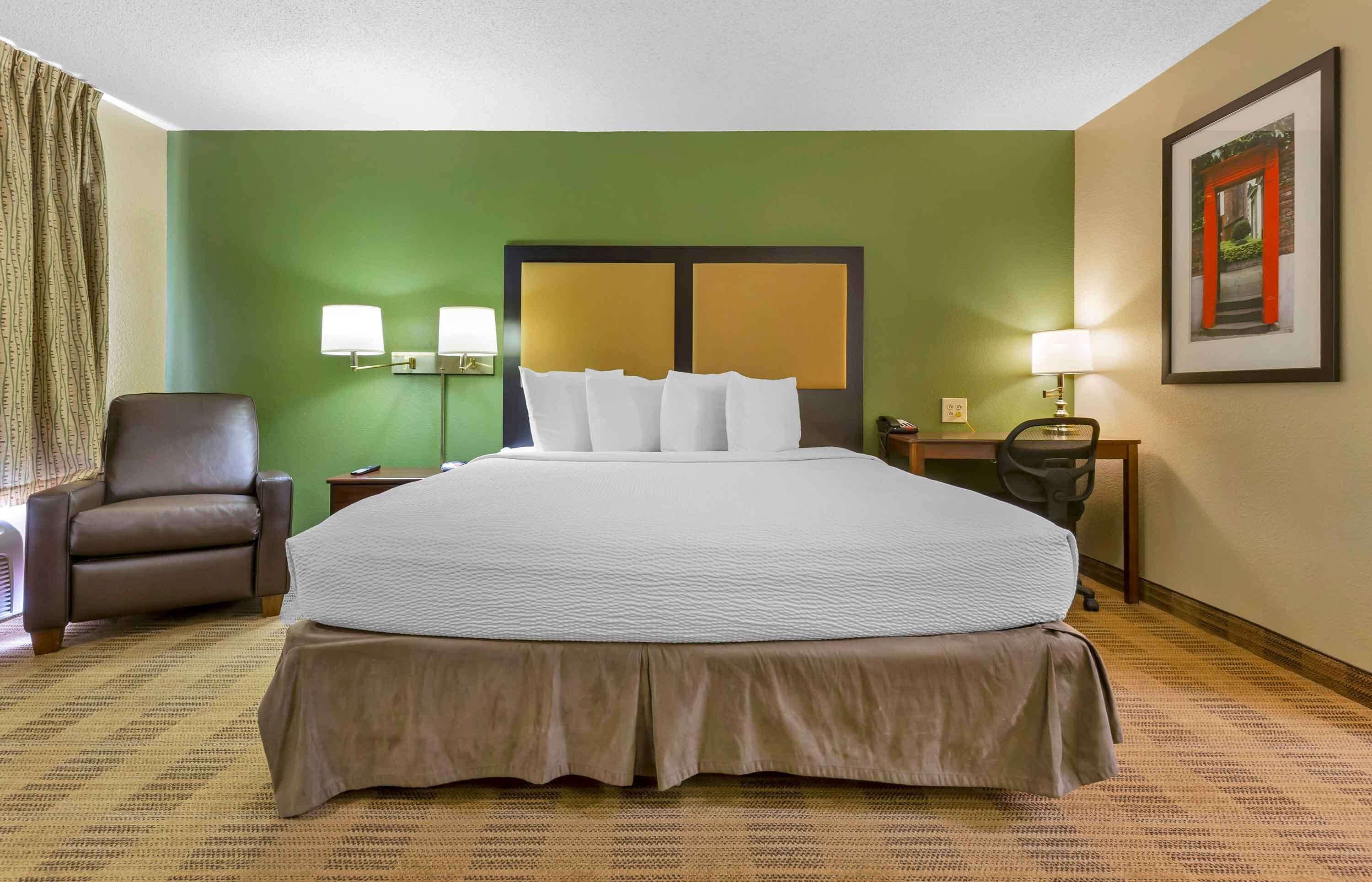 Une chambre d’hôtel avec un grand lit, une chaise, deux tables de chevet avec des lampes et un tableau encadré au mur.