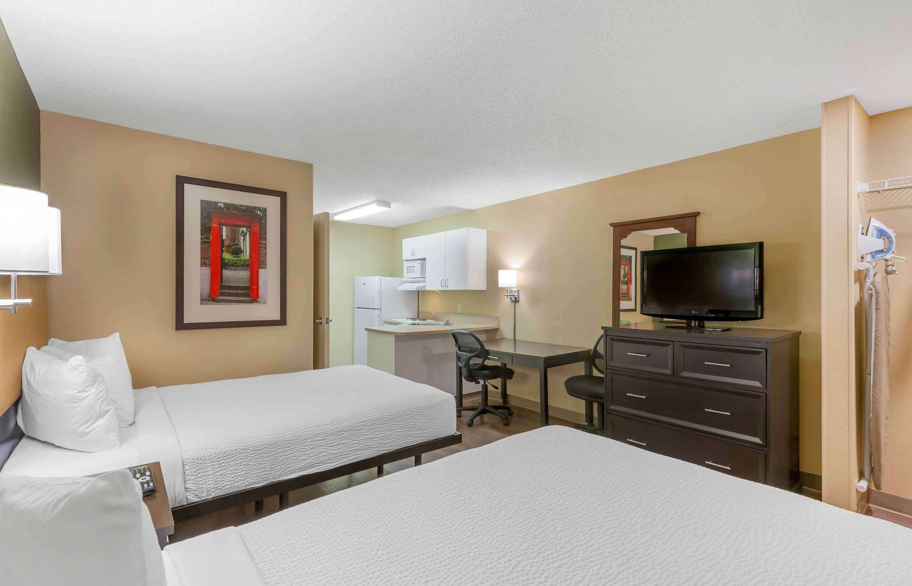A hotel room with two beds, a desk, a television, and a kitchenette.
