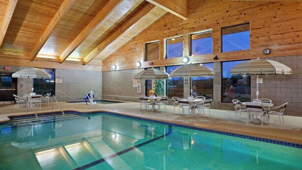 An indoor swimming pool with wooden ceiling beams, tiled walls, and multiple lounge areas with umbrellas. - AmericInn by Wyndham Chanhassen (Chanhassen)