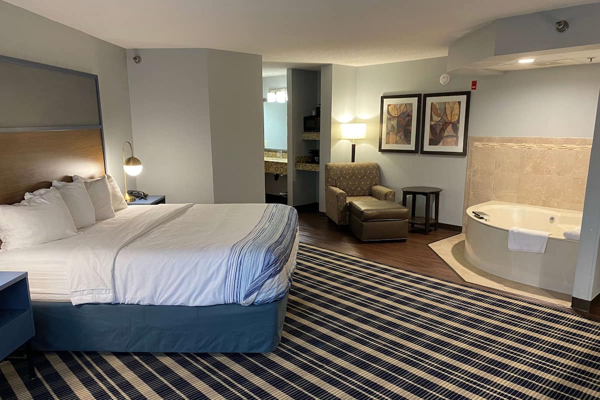 Ein Hotelzimmer mit einem großen Bett, einem Sessel, einem kleinen Tisch, einer Lampe, einem Badezimmer mit Badewanne und Bildern an der Wand.