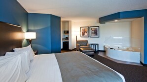 A hotel room with a large bed, a sitting area with a chair and a small table, a bathtub, and two framed pictures on the wall. - AmericInn by Wyndham Chanhassen (Chanhassen)