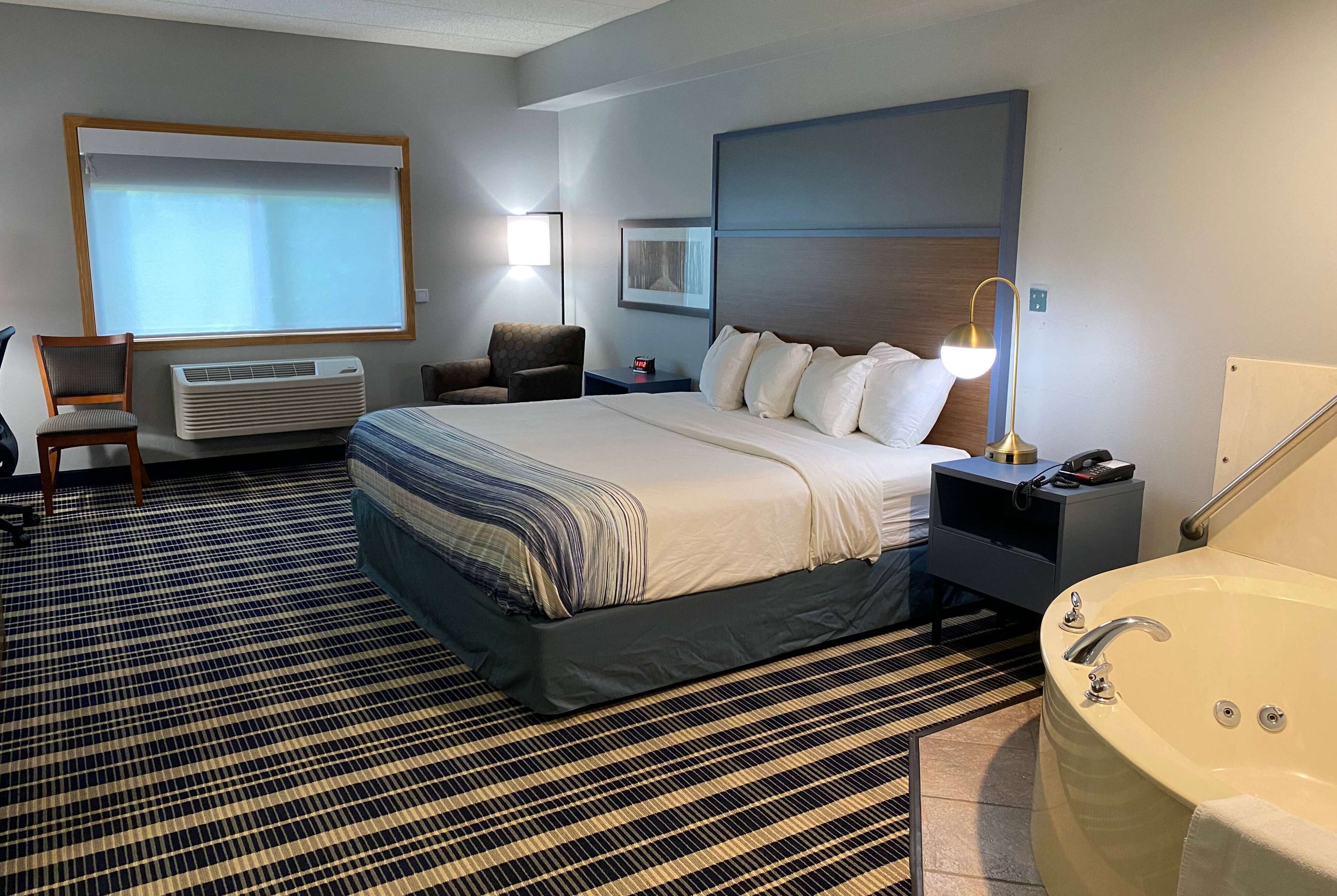 A hotel room with a large bed, a sitting area with a chair, a desk with a lamp, a large window with blinds, and a bathtub.