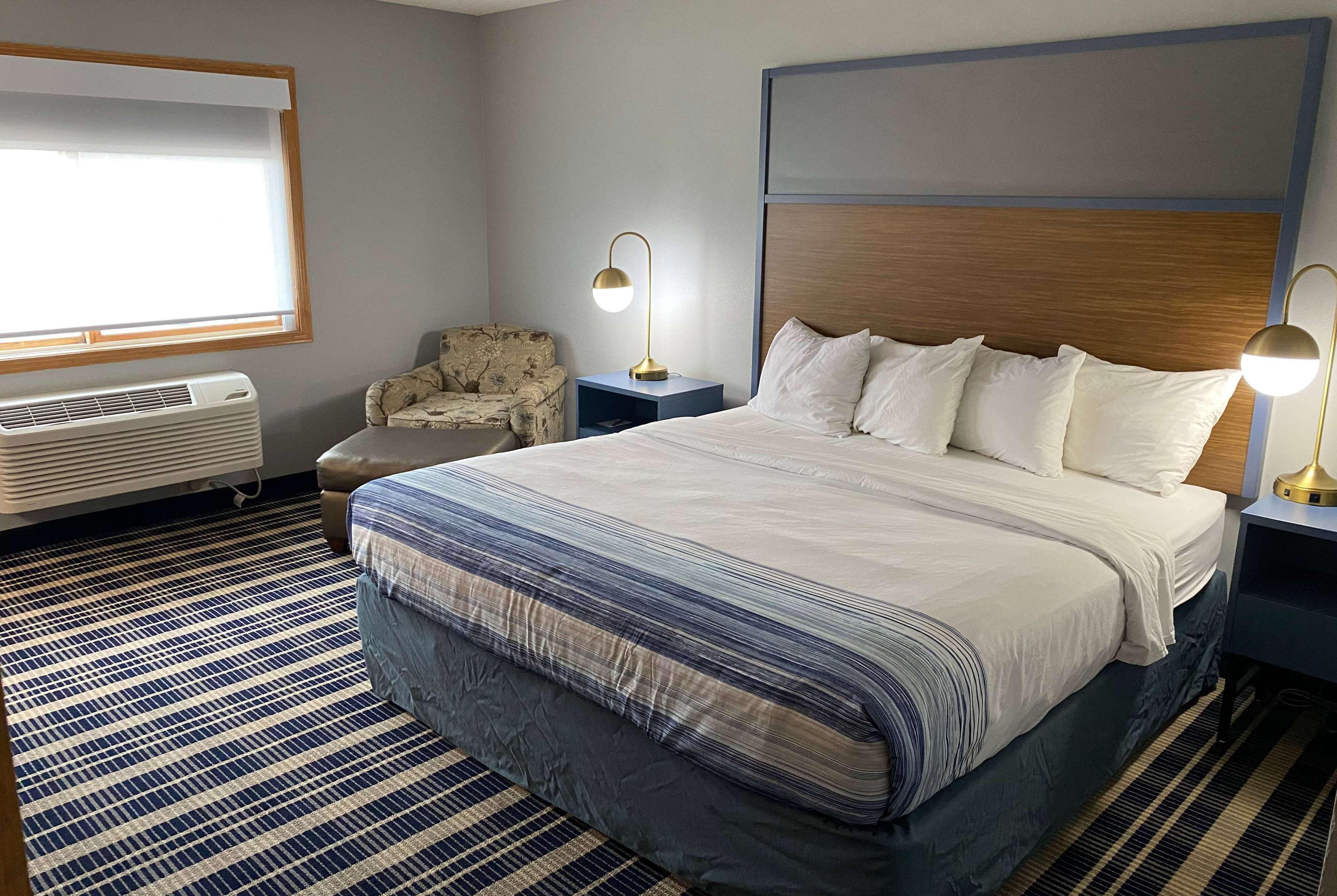 Ein Hotelzimmer mit einem großen Bett, zwei Nachttischen mit Lampen, einem Sessel, einem Fenster mit Jalousien und einer Wand-Klimaanlage.