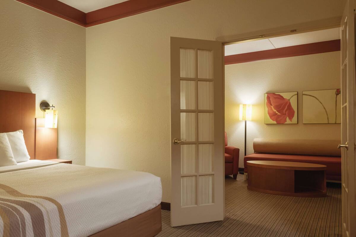 A hotel room with a bed, a seating area with a bench, a door, and a wall with a painting.