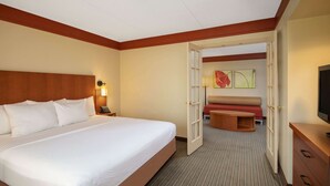 A hotel room with a large bed, a desk with a lamp, a television, and a door leading to a seating area.