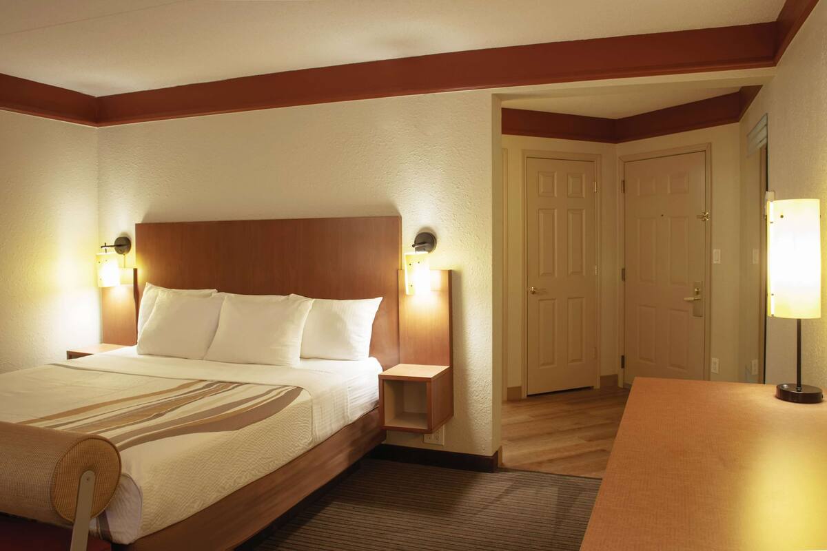A hotel room with a large bed, bedside lamps, a wooden headboard, and a door leading to another room.