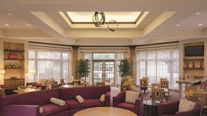 A spacious living area with a large sofa, armchairs, and a round coffee table. There are multiple dining tables and chairs, and a television mounted on the wall.