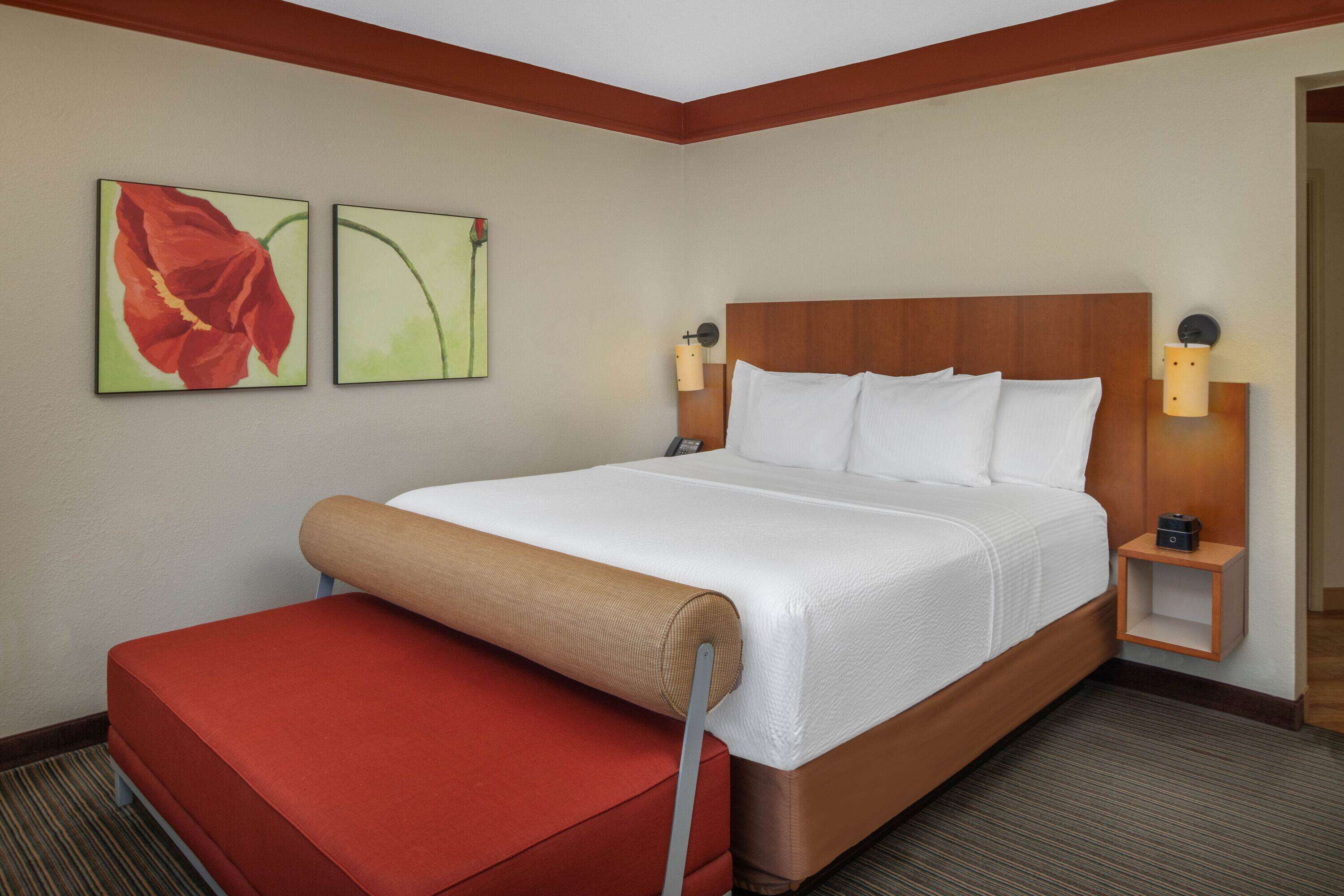 Una habitación de hotel con una cama grande, un banco rojo, dos lámparas de pared y dos cuadros.