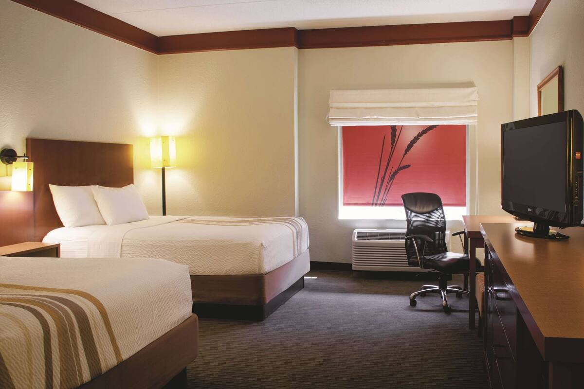 A hotel room with two beds, a desk with a television, a chair, and a window with a view of tall grasses.