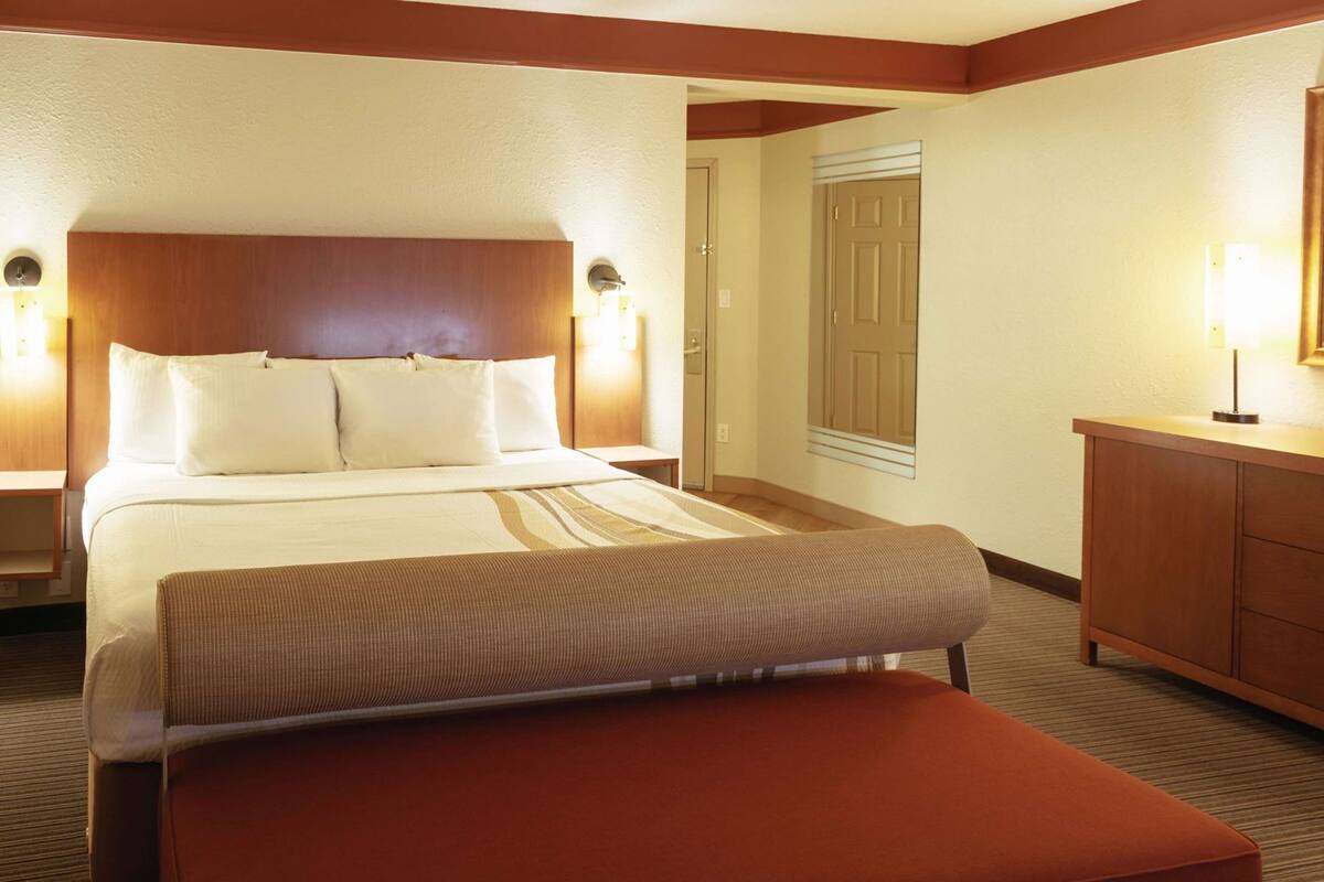 A hotel room with a bed, bedside tables, a wooden headboard, a bench, a dresser, and a door.