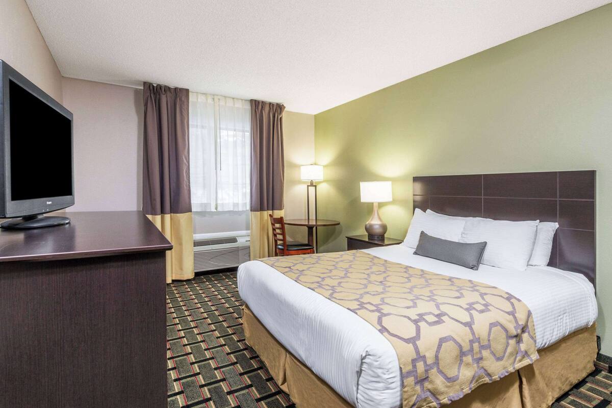Habitación de hotel con cama, escritorio con televisor, silla, dos lámparas y ventana con cortinas.
