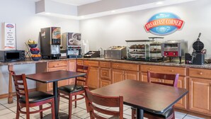 Una barra de desayuno con un letrero que dice "Baymont Breakfast Corner", un microondas, una máquina de café y un fabricante de gofres. Hay sillas de madera y una mesa frente a la barra.