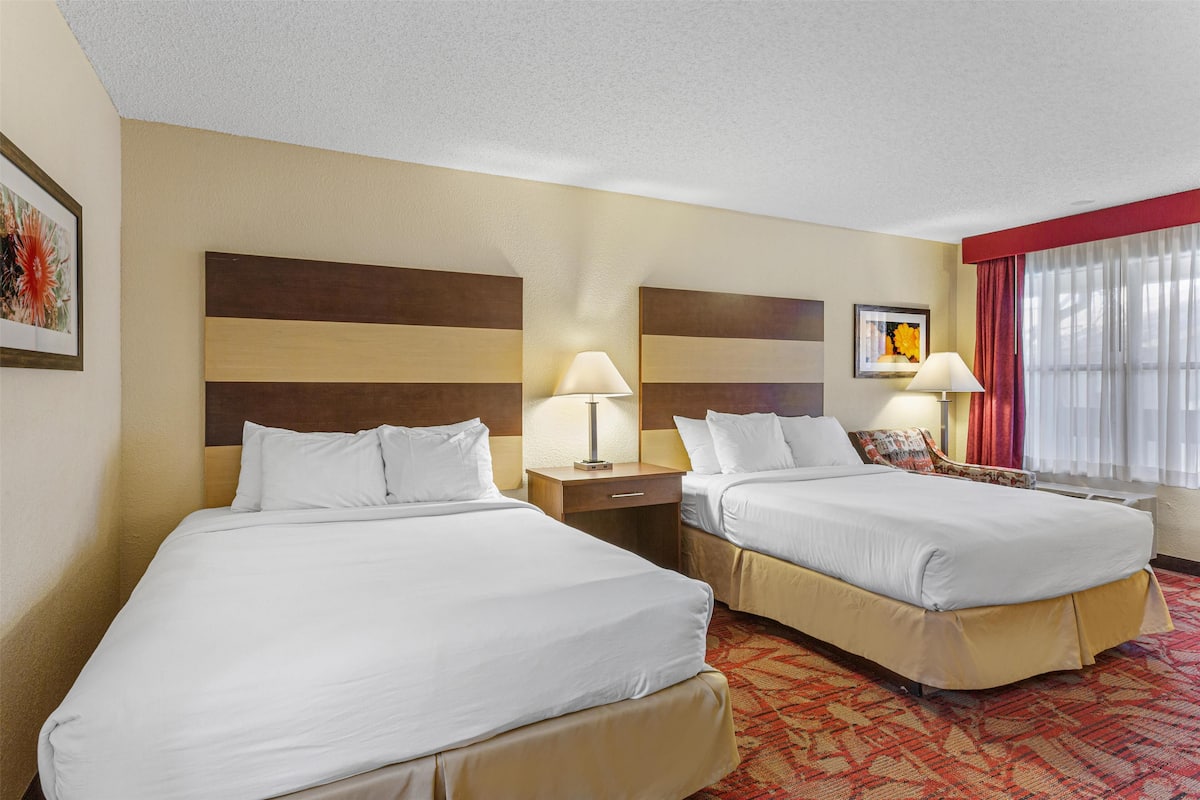 A hotel room with two beds, a nightstand with a lamp, a chair, and a window with curtains.