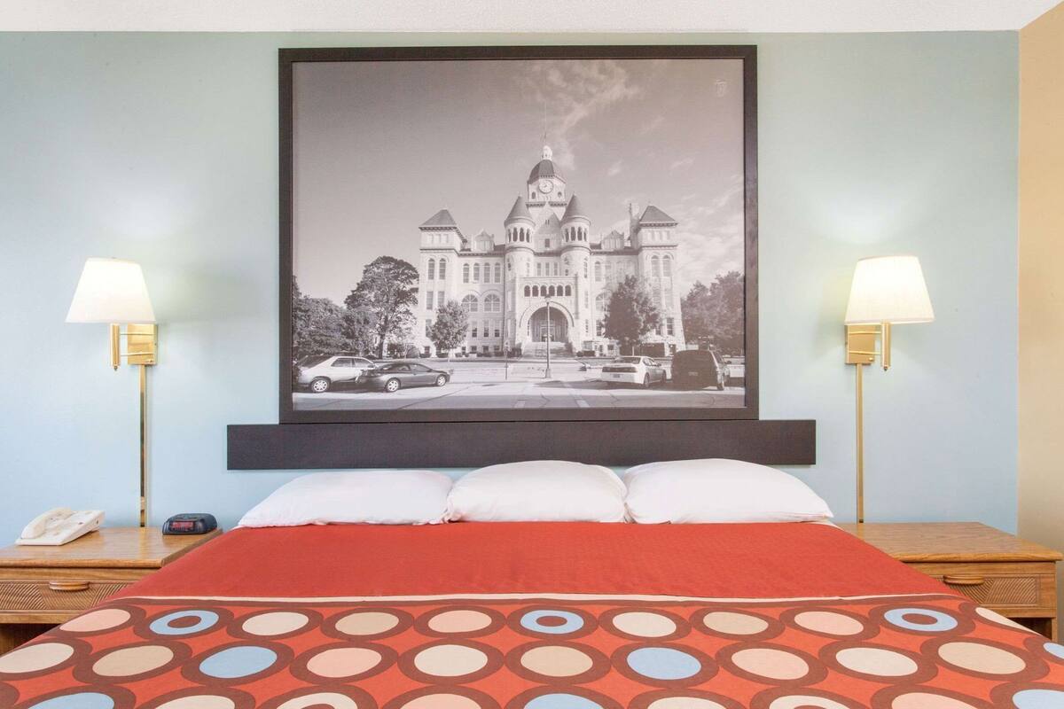 A hotel room with a bed, two bedside lamps, a framed picture of a building, and a telephone on the nightstand.
