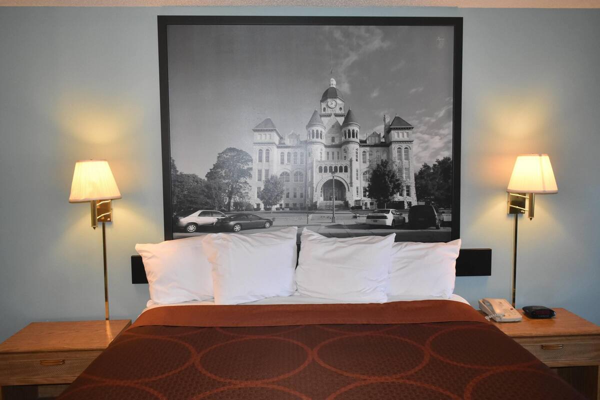 A hotel room with a bed, two bedside lamps, a framed picture of a building, and a nightstand with a phone.