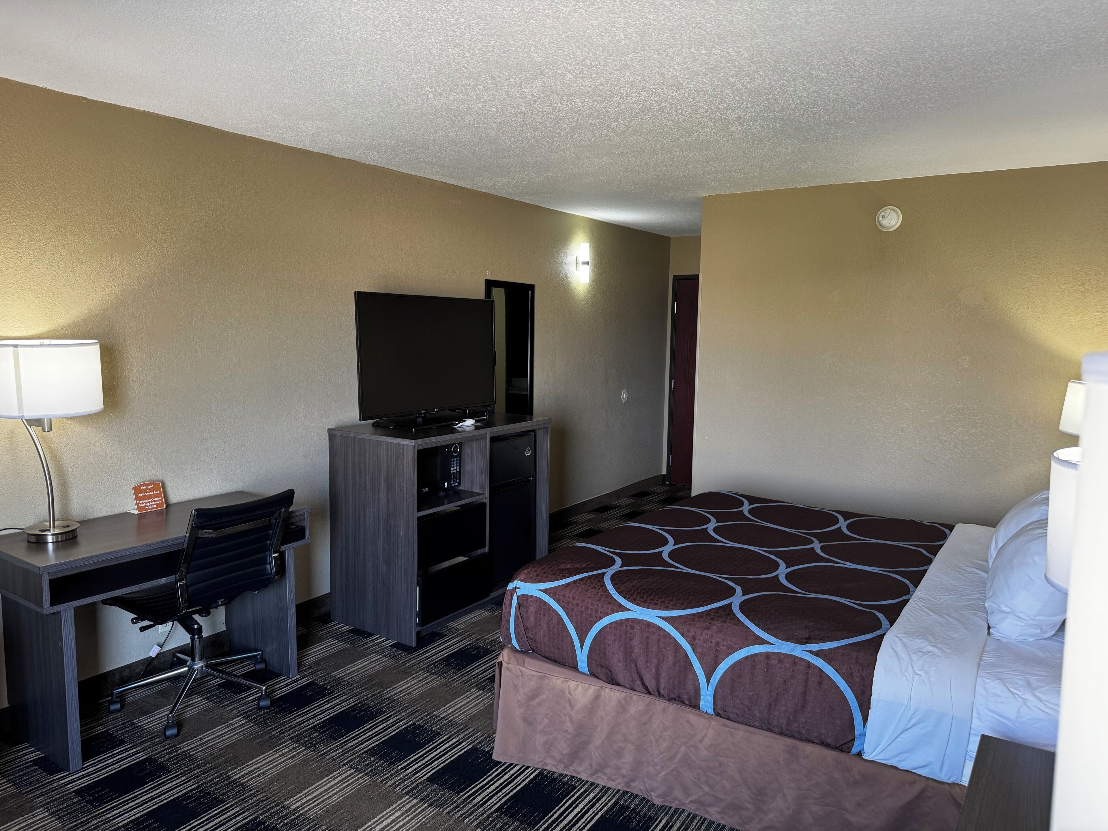 A hotel room with a bed, desk, TV, and a small refrigerator.