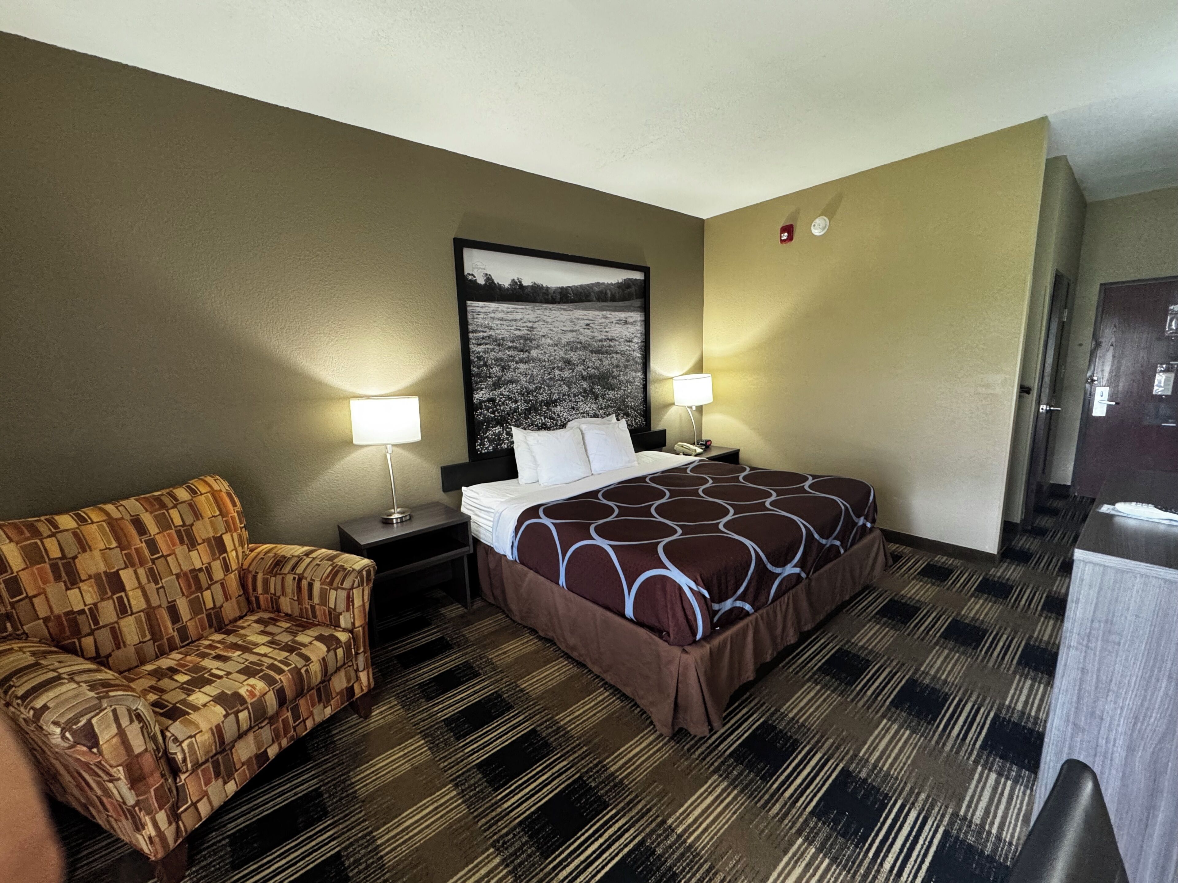 Una habitación de hotel con una cama, dos mesitas de noche, una silla, un cuadro grande en la pared y una puerta.