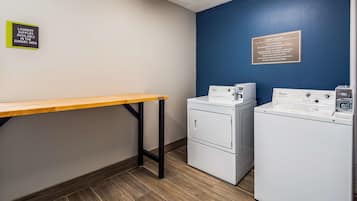 Zona de bugaderia amb dues rentadores Whirlpool, una taula i indicadors que mostren servei de bugaderia per a clients i disponibilitat de productes de neteja.
