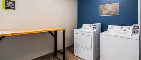 A laundry area with two Whirlpool washers, a table, and signs indicating guest laundry and laundry supplies availability.