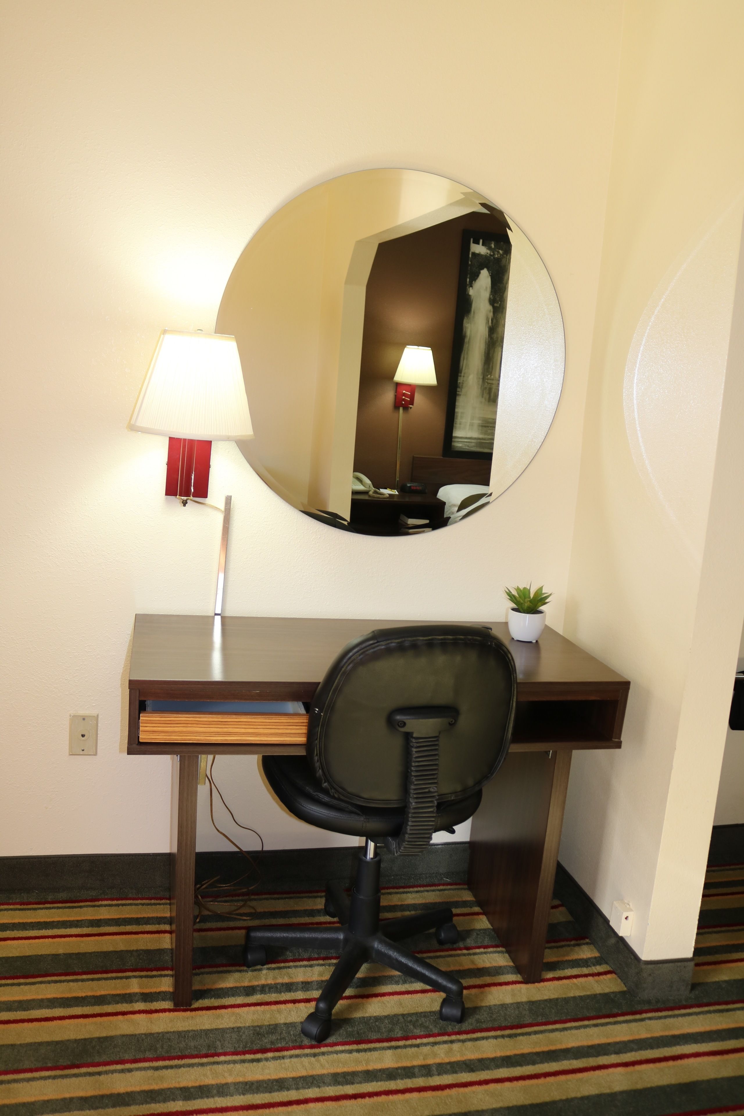 A hotel room with a wooden desk, a black office chair, a round mirror, a lamp, and a small potted plant.