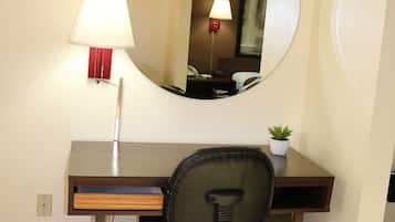 A hotel room with a wooden desk, a black office chair, a round mirror, a lamp, and a small potted plant.