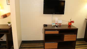 A hotel room with a flat-screen TV mounted on the wall, a wooden desk with drawers, a small potted plant, and a striped carpet.