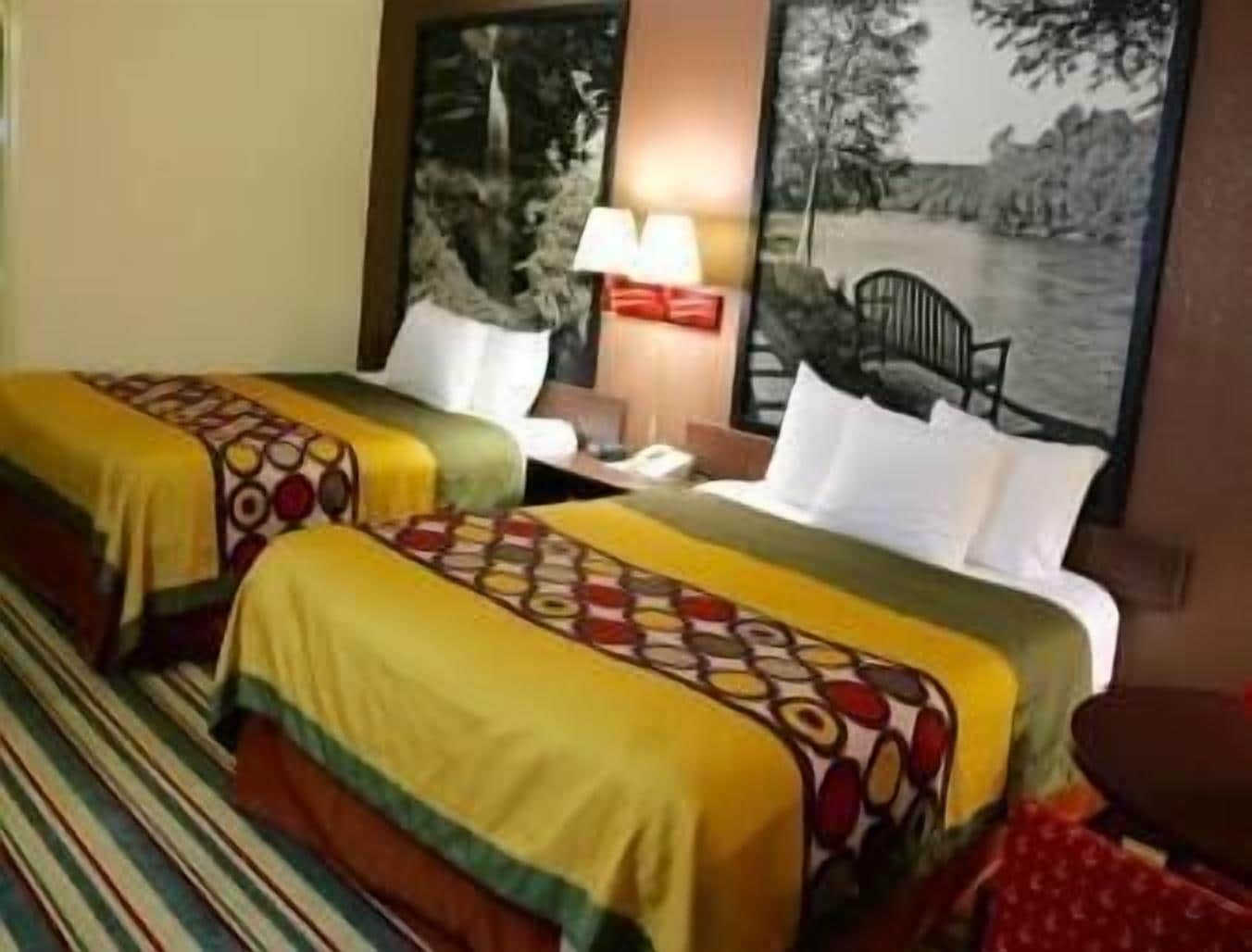 A hotel room with two beds, each with patterned bedspreads, a nightstand with a lamp, and a painting of a landscape on the wall.