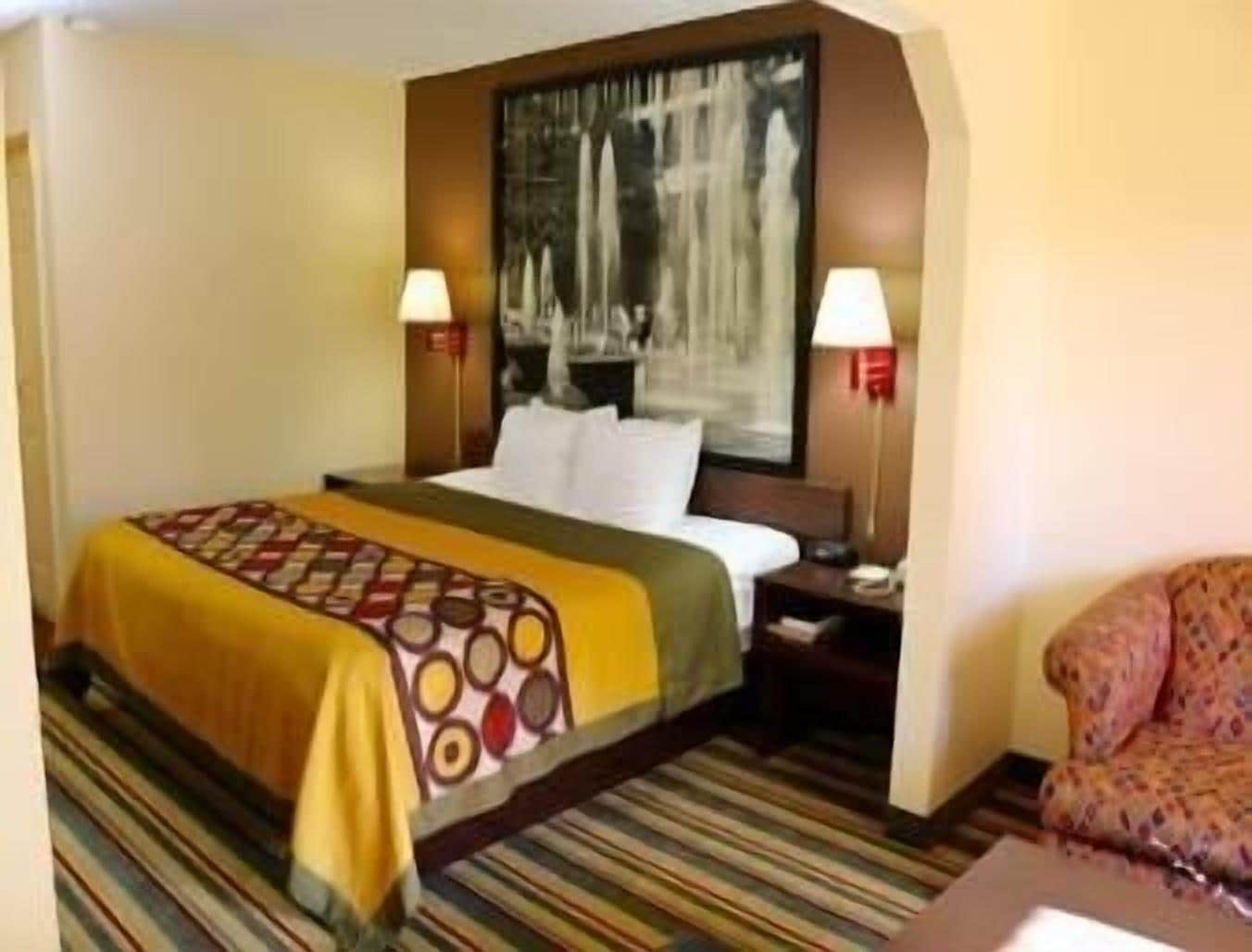 Habitación de hotel con una cama, dos lámparas de noche, una mesita de noche, una alfombra estampada y un cuadro en la pared.