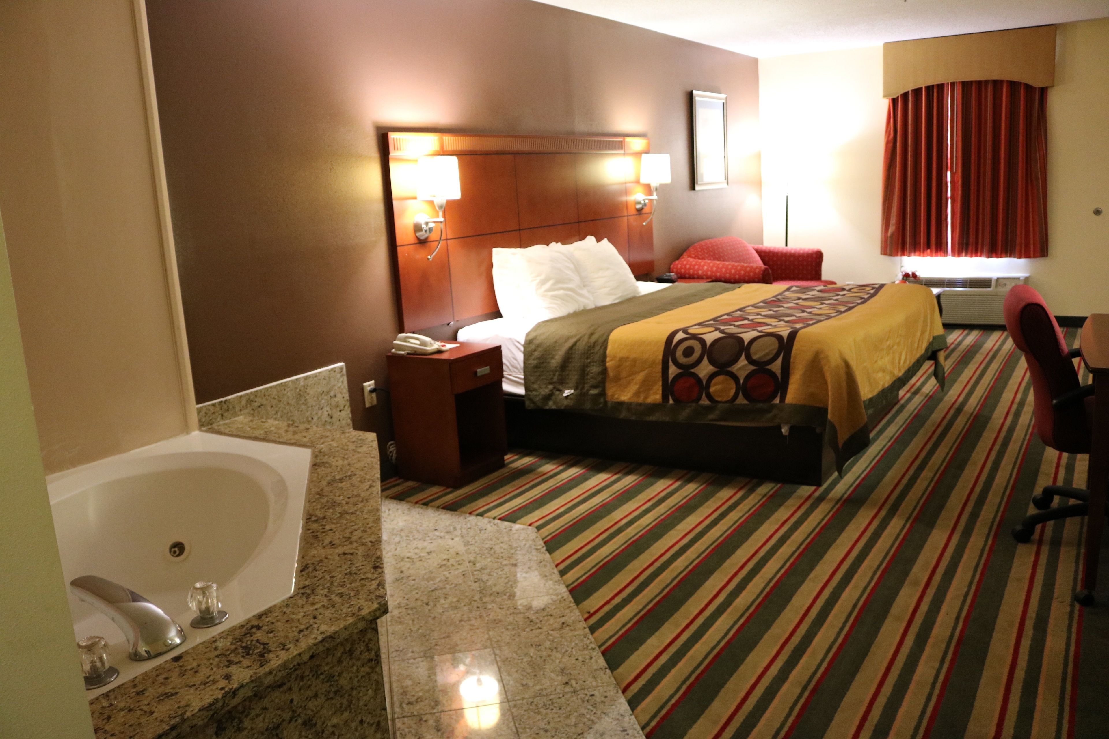 Habitación de hotel con una cama grande, una zona de estar con una silla, un baño con lavamanos y espejo, y una ventana con cortinas.