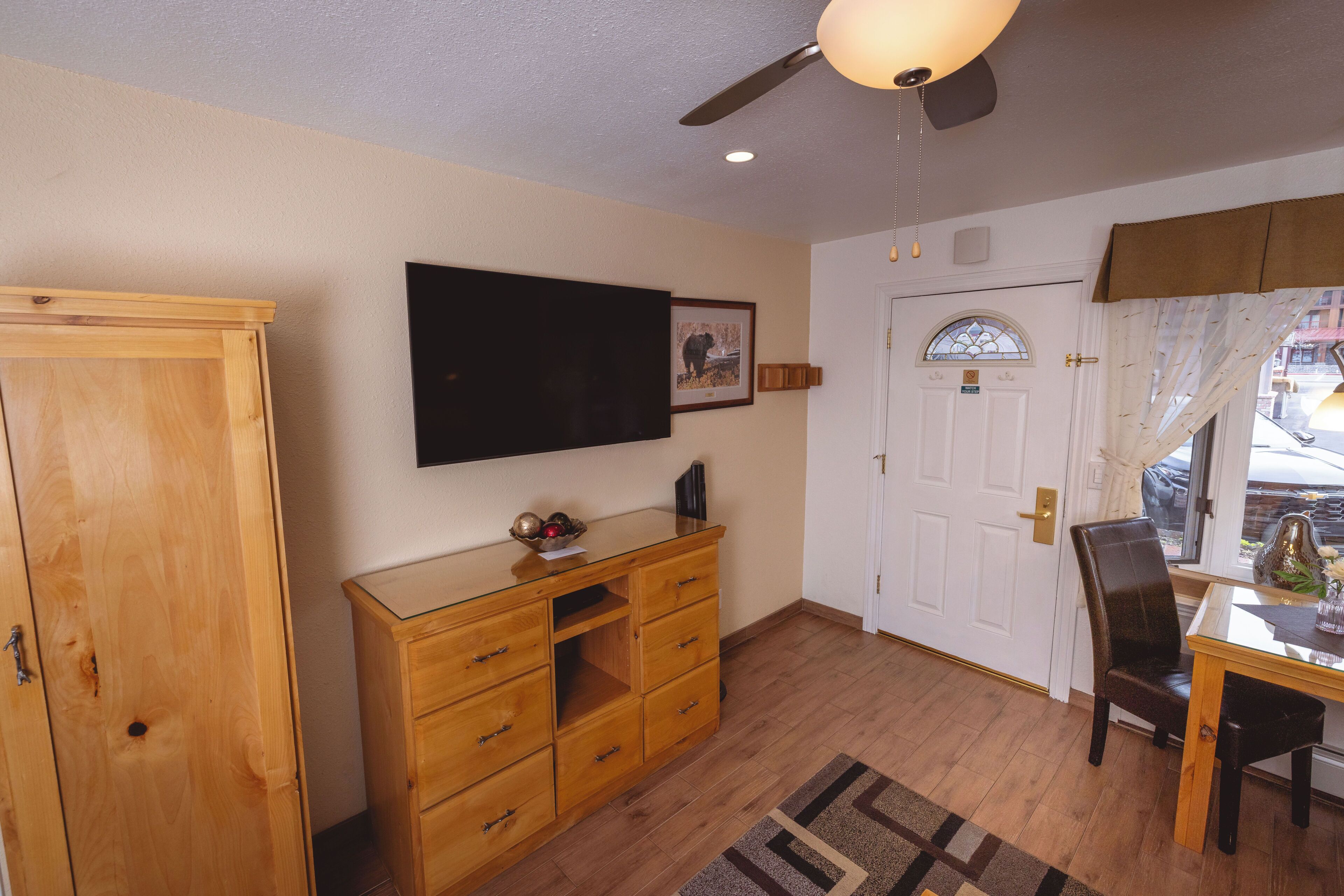 A room with a wooden cabinet, a flat-screen TV, a dining table with chairs, a ceiling fan, and a door with a window.