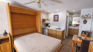 A bedroom with a wooden bed, a ceiling fan, a kitchenette, and a dining area.
