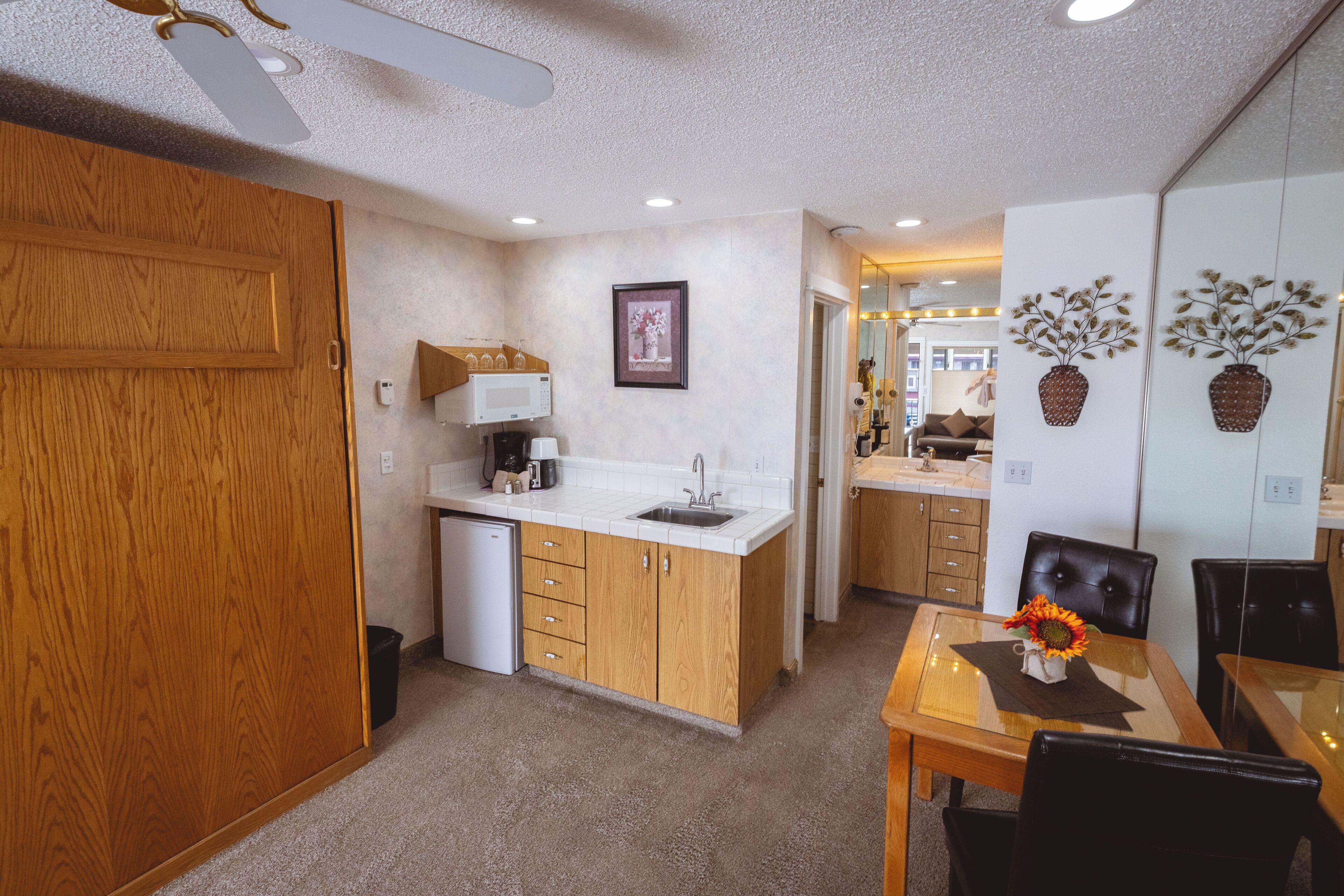 A compact kitchen with wooden cabinets, a microwave, and a sink. There is a dining table with chairs and a vase with flowers.