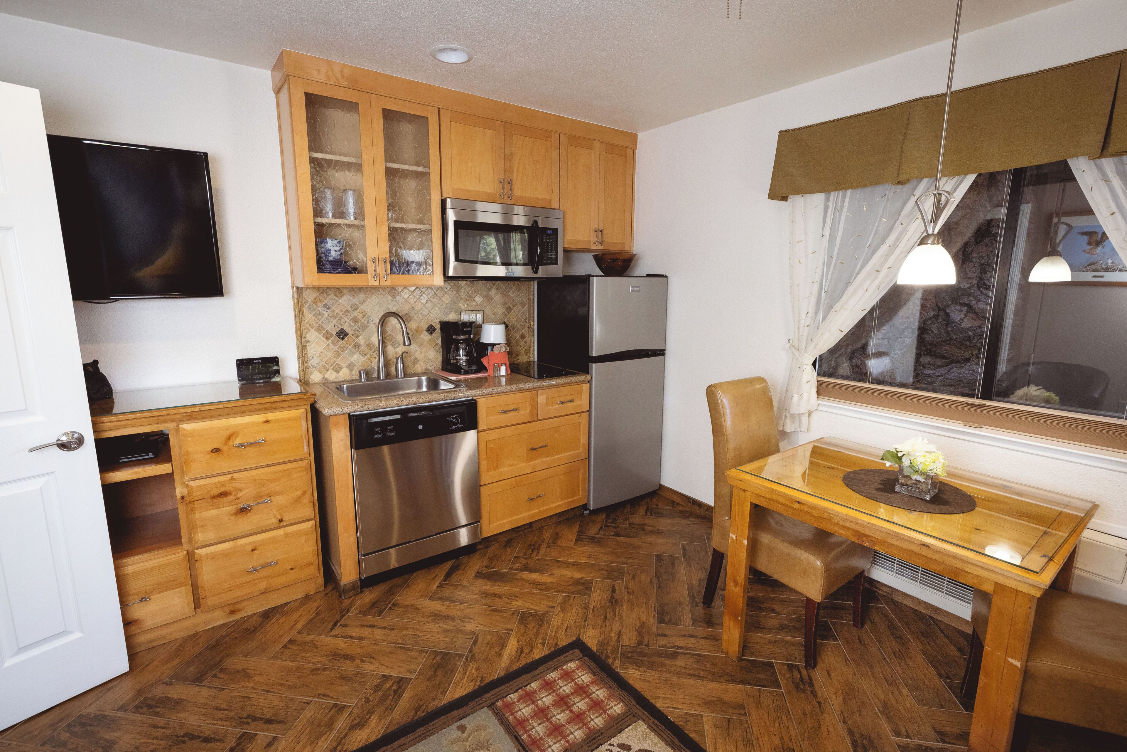 Cocina pequeña con armarios de madera, microondas, refrigerador, lavavajillas y una mesa de comedor con una silla.