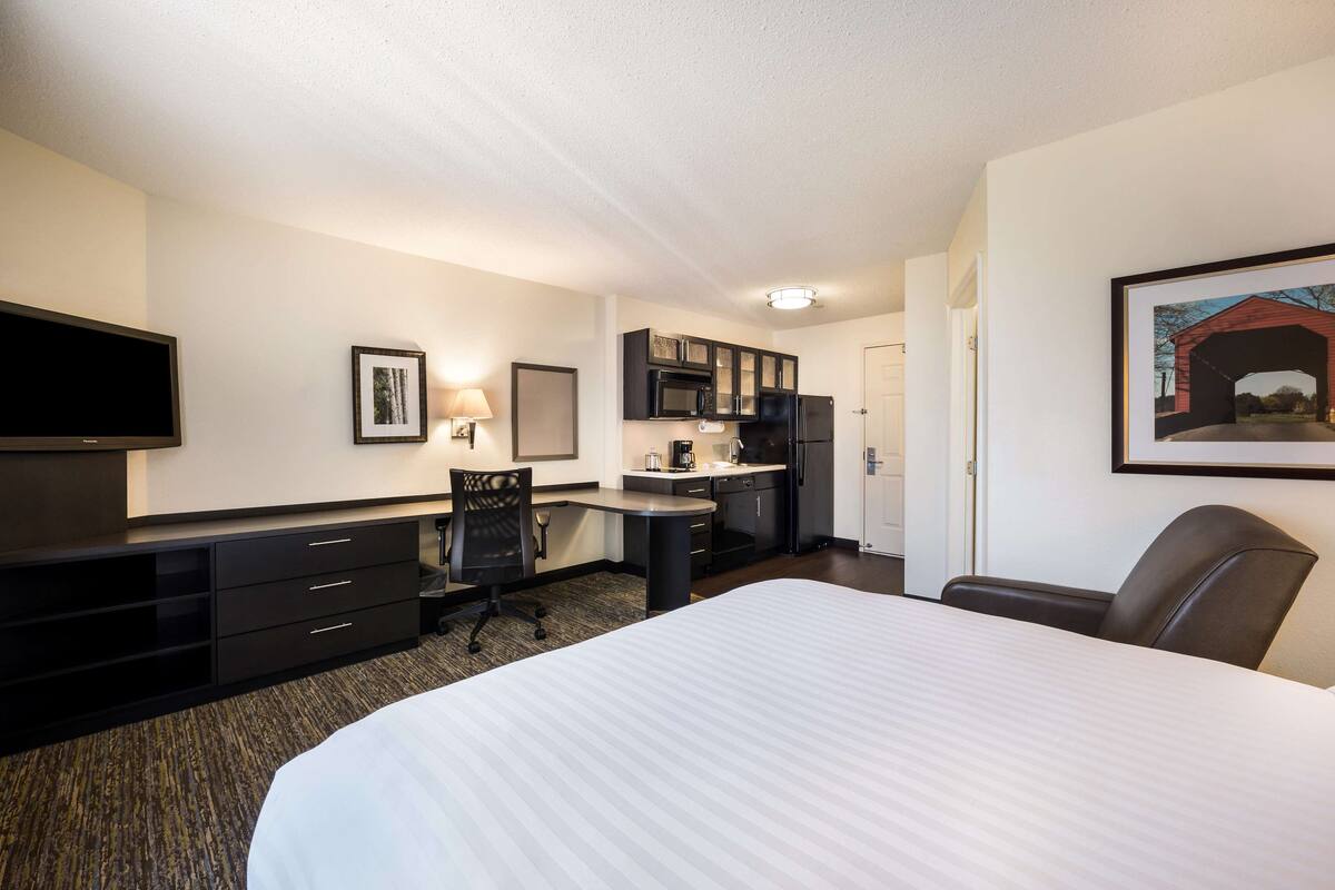 Habitación de hotel con cama, escritorio con silla, kitchenette y televisor.