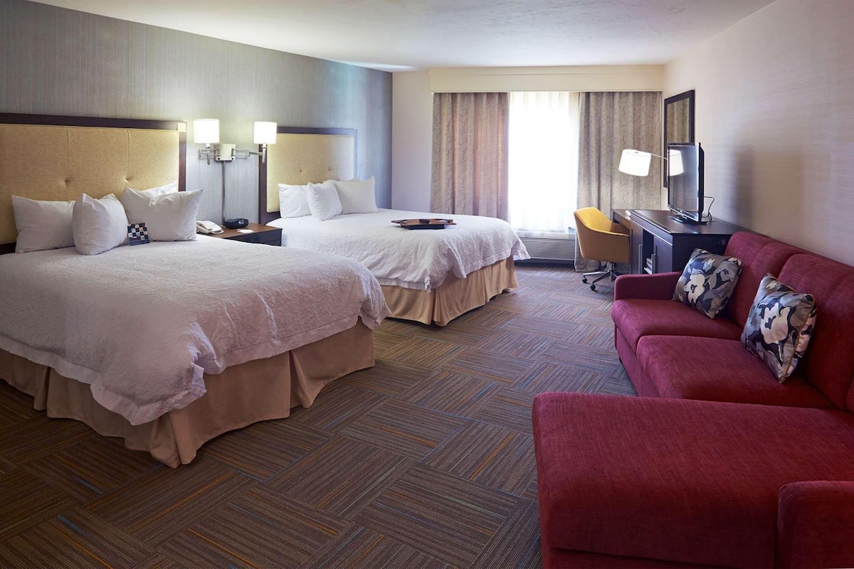 A hotel room with two beds, a red sofa, a desk with a lamp, and a television.